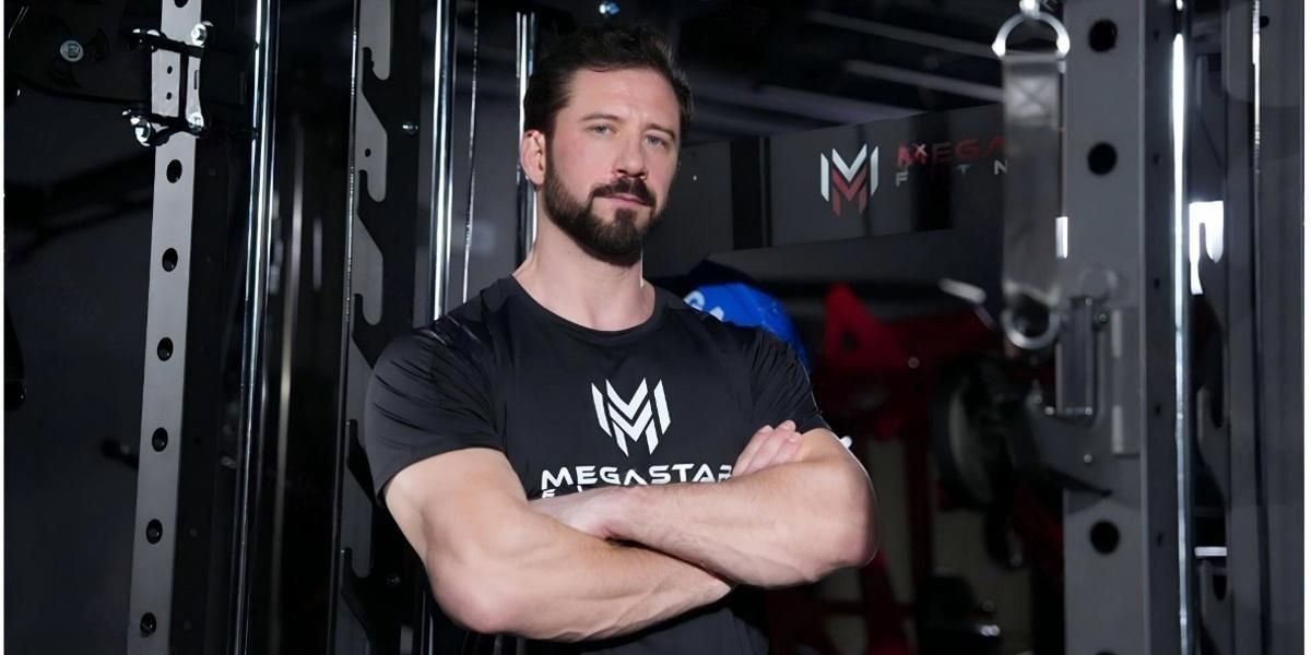 A man in a Megastar Fitness t-shirt standing with arms crossed in front of gym equipment, promoting strength and fitness in a beginner’s guide to workout recovery.