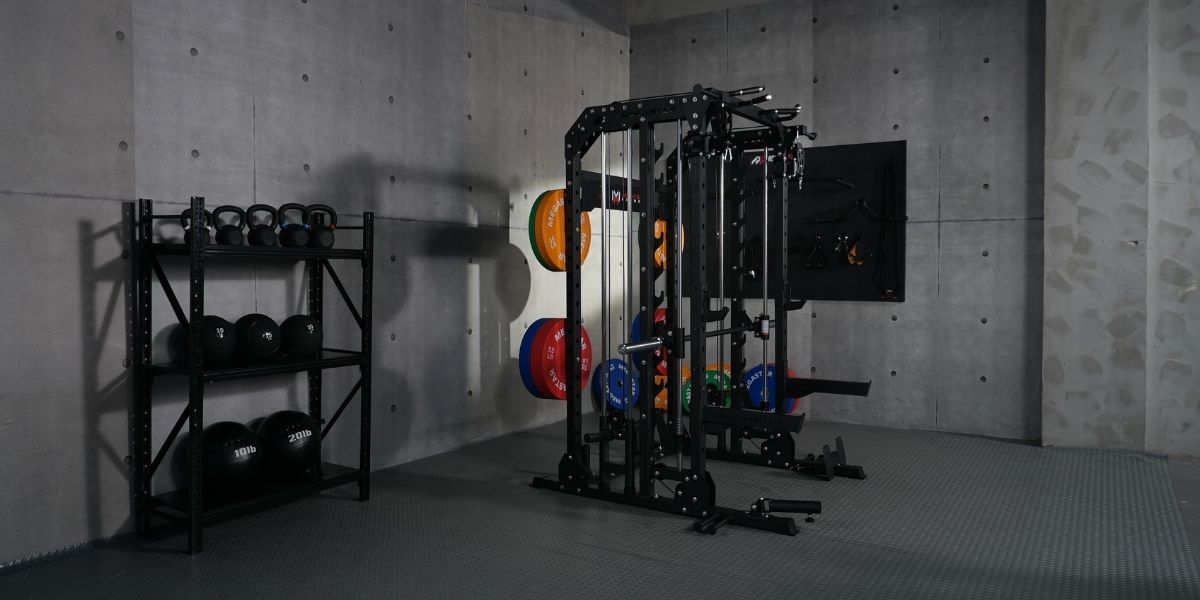 Angled view of a smith machine with cable pulleys and colorful bumper plates in a modern concrete gym, with a kettlebell and slam ball rack nearby.