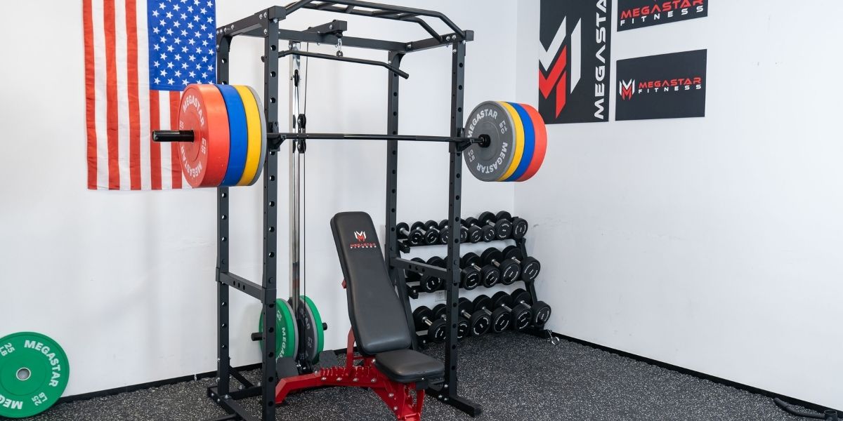 Home gym setup featuring a squat rack with colorful weight plates, a bench, and dumbbell racks, with a USA flag and Megastar Fitness logos on the wall.