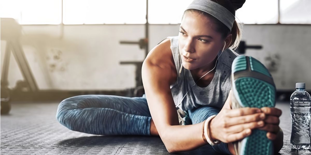 Woman stretching on a gym floor during training, doing a seated hamstring stretch with headphones on and a water bottle nearby.